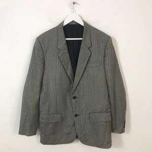 May include: A grey blazer with a subtle herringbone pattern, two front pockets, and a black interior. The blazer is hung on a white hanger against a white background. It has two buttons and a notched lapel.