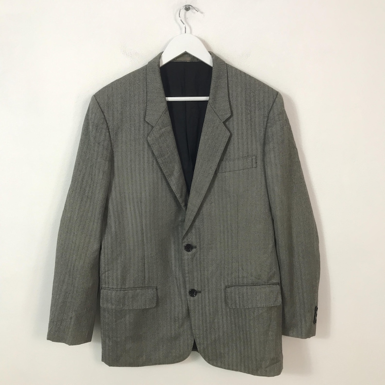 May include: A grey blazer with a subtle herringbone pattern, two front pockets, and a black interior. The blazer is hung on a white hanger against a white background. It has two buttons and a notched lapel.