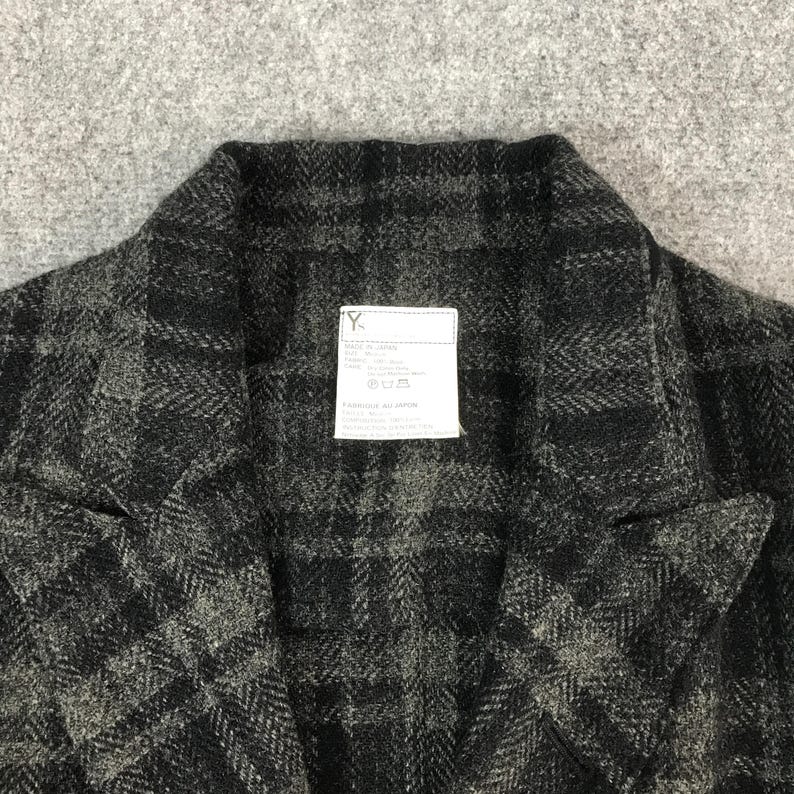 May include: A close-up shot of a dark grey and black checked jacket. The jacket features a notched collar and a small white label with text. The fabric has a textured finish.