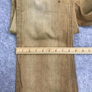 May include: Close-up of a pair of brown denim jeans with a ruler measuring 28 cm. The jeans have a faded, distressed look.