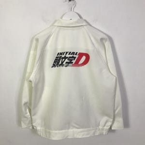 Puede incluir: Chaqueta blanca con cuello y gráfico negro, rojo y blanco en la espalda que dice "INITIAL D". La chaqueta tiene cierre de cremallera y puños y dobladillo elásticos. La chaqueta está colgada de una percha blanca.