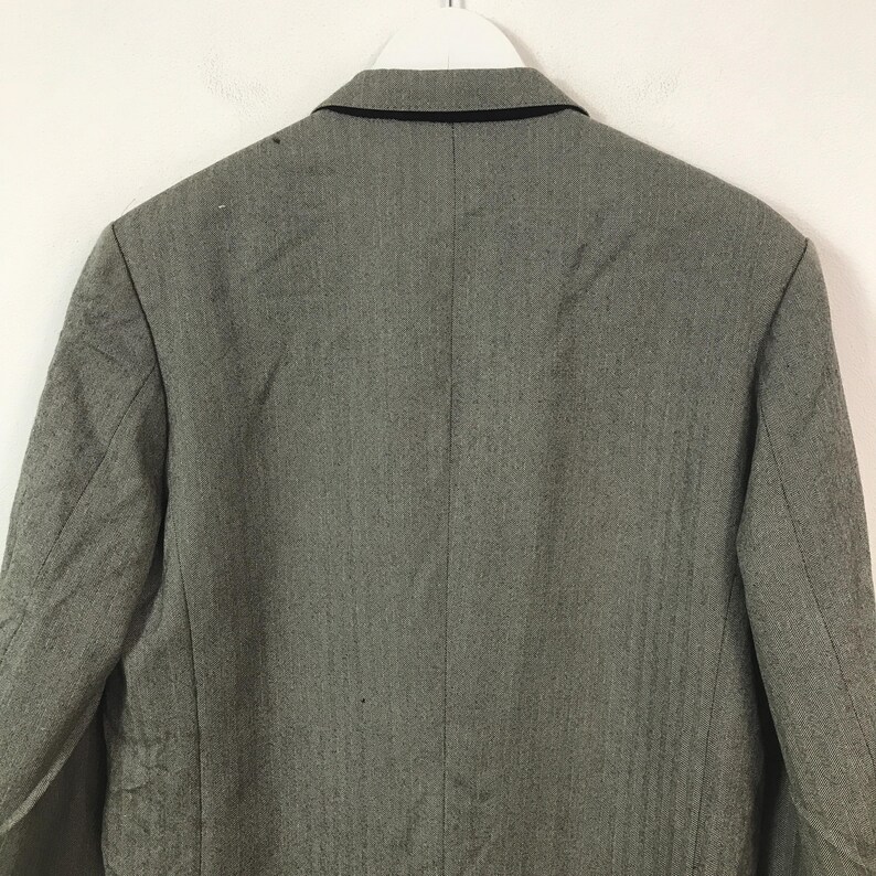 May include: A grey blazer with a black collar, suspended from a white hanger. The rear view of the blazer reveals its tailored design and fabric texture. The blazer has a classic, professional style.