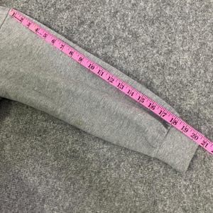 May include: A gray sweatshirt sleeve is measured with a pink measuring tape. The tape shows measurements up to 21 inches. The sleeve is on a gray textured surface.