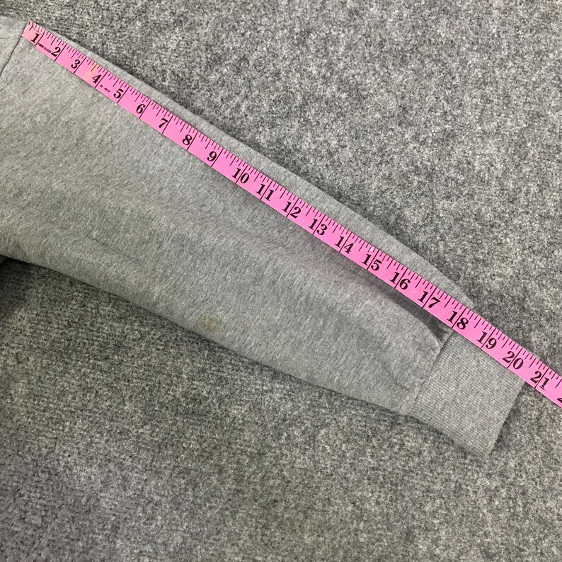 May include: A gray sweatshirt sleeve is measured with a pink measuring tape. The tape shows measurements up to 21 inches. The sleeve is on a gray textured surface.