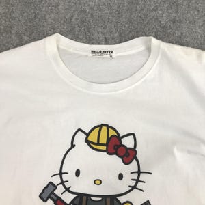 May include: White t-shirt featuring a cartoon image of Hello Kitty wearing a yellow hard hat and holding a hammer. The tag reads "HELLO KITTY".