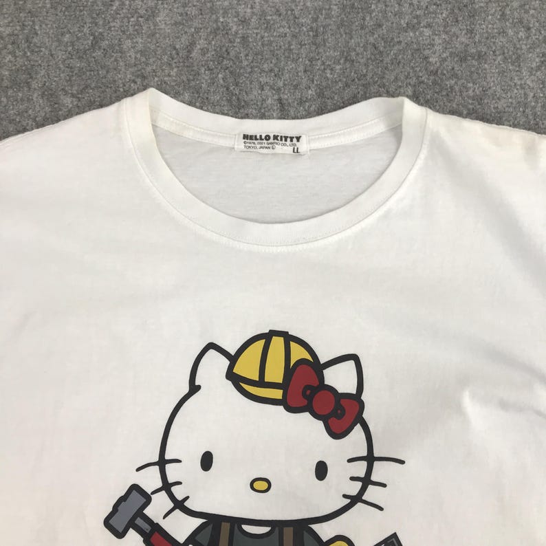 May include: White t-shirt featuring a cartoon image of Hello Kitty wearing a yellow hard hat and holding a hammer. The tag reads "HELLO KITTY".