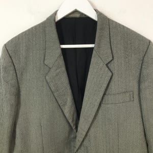 May include: A grey blazer with a black lining, suspended from a white hanger. The jacket features a notched lapel, a chest pocket, and a two-button fastening. The material has a subtle textured design.