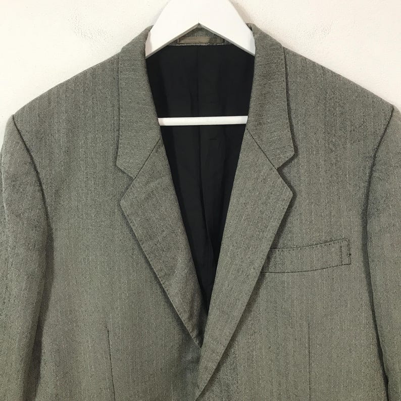 May include: A grey blazer with a black lining, suspended from a white hanger. The jacket features a notched lapel, a chest pocket, and a two-button fastening. The material has a subtle textured design.