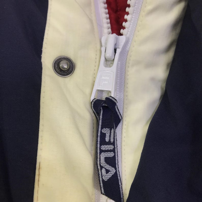 May include: Close-up of a jacket zipper with a white zipper pull and a navy blue fabric pull tab with the brand name "FILA" in white. The jacket has a cream-colored panel, navy blue side panel, and a red interior.