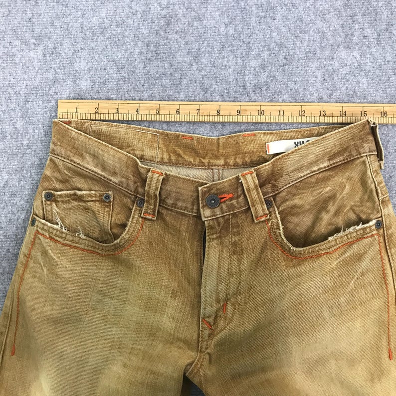 May include: A pair of brown denim jeans with orange stitching. The jeans have a faded look and a worn-in appearance.