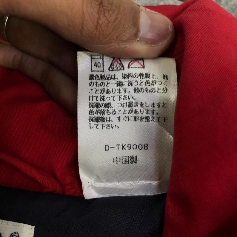 May include: A close-up of a clothing tag with Japanese text and the number 40. The tag is white with black text and is held against a red fabric. The text includes the product code D-TK9008 and the words "Made in China".