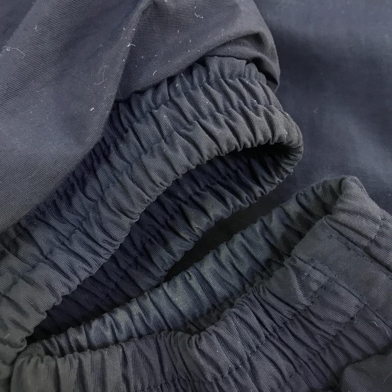 May include: Close-up of navy blue fabric, showcasing the elasticized cuffs of a garment. The fabric has a slightly textured appearance, with visible stitching and gathered details. The image highlights the design and construction of the clothing item.