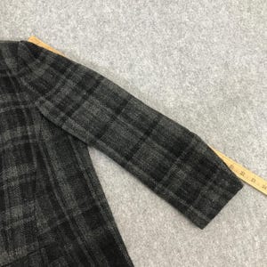 May include: A close-up of a grey and black plaid blazer sleeve. The fabric has a textured appearance with a classic plaid pattern. A wooden ruler is visible along the edge of the sleeve.