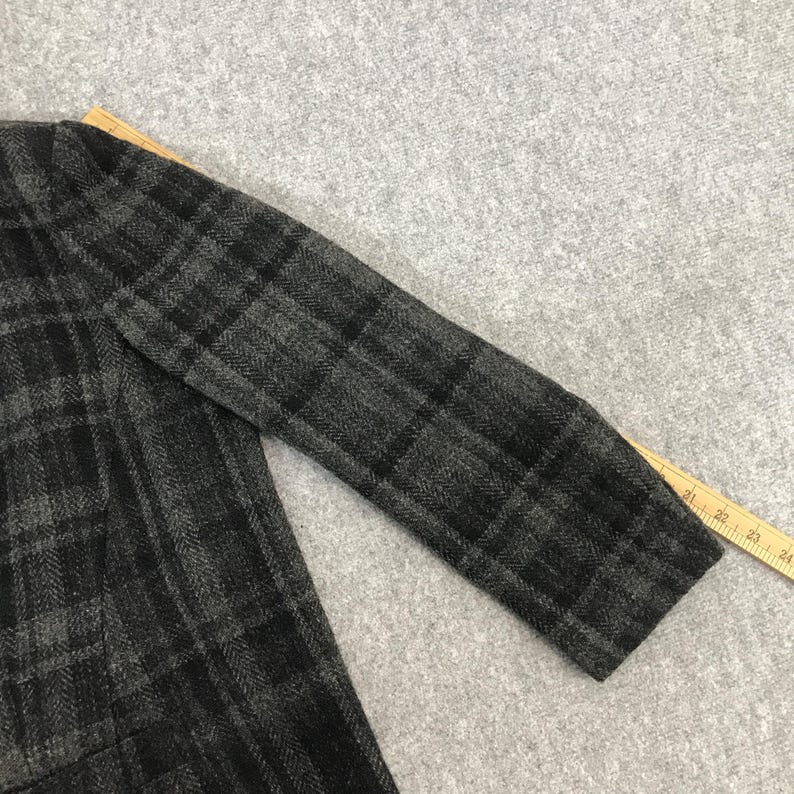 May include: A close-up of a grey and black plaid blazer sleeve. The fabric has a textured appearance with a classic plaid pattern. A wooden ruler is visible along the edge of the sleeve.