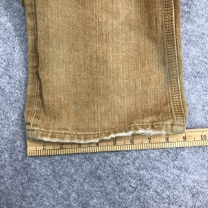 May include: Close-up of the bottom edge of a pair of brown denim trousers. The fabric is frayed and the trousers are measured with a ruler.
