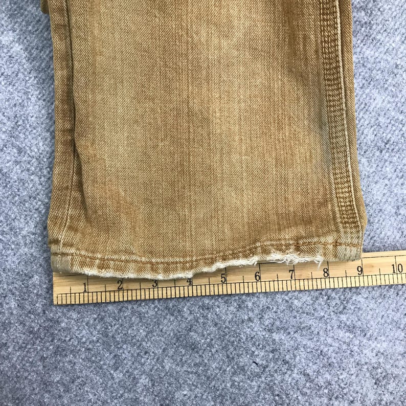 May include: Close-up of the bottom edge of a pair of brown denim trousers. The fabric is frayed and the trousers are measured with a ruler.