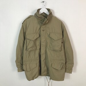 May include: A khaki-coloured military-style jacket with multiple front pockets and a high collar. The jacket is hanging on a white hanger against a white background. It features a button-up front and a drawstring at the bottom.