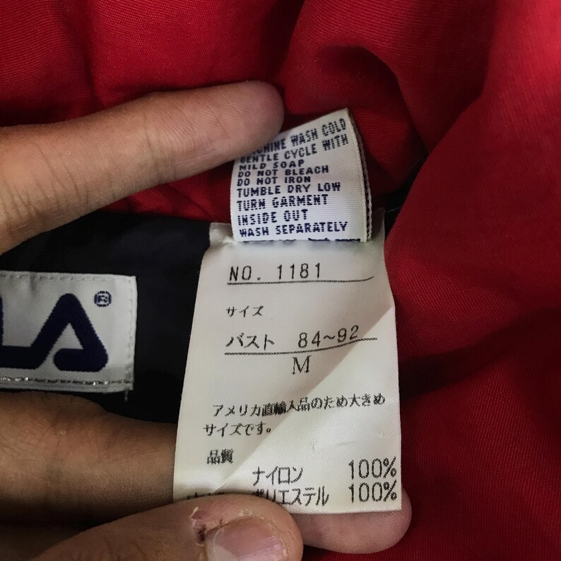 May include: Close-up of a garment tag with the Fila logo. The tag includes washing instructions and size information. The text reads "NO. 1181", "サイズ", "バスト 84~92", and "M". The fabric composition is 100% nylon and 100% polyester.