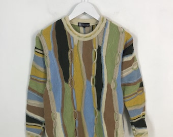 May include: A colorful, patterned sweater with a crew neck. The sweater features vertical stripes in various colors, including blue, green, yellow, and brown. The sleeves and hem have a cream-colored trim. The sweater is hanging on a white hanger.