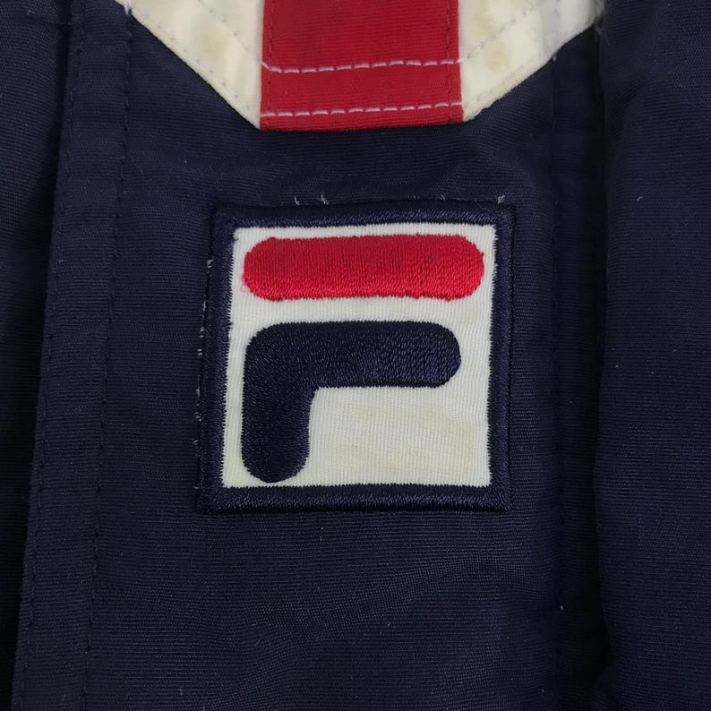 May include: Close-up of a navy blue jacket with a white, red, and navy blue design. The jacket features a square patch with the brand's logo in red, white, and navy blue.