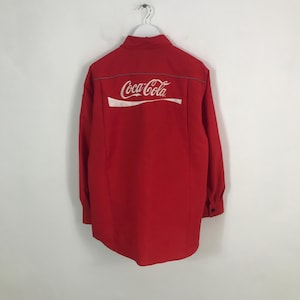 Può includere: Una camicia rossa brillante a maniche lunghe con il logo Coca-Cola stampato in bianco sul retro. La camicia ha un'abbottonatura frontale e un colletto. Il logo Coca-Cola è una scritta stilizzata con un'onda bianca sotto.