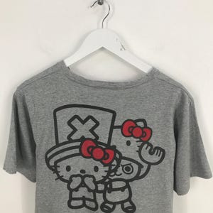 Pu&ograve; includere: T-shirt grigio con una grafica di due personaggi dei cartoni animati con fiocchi rossi. Un personaggio indossa un cappello con un simbolo a croce. L'altro personaggio ha un fiocco rosso e sta salutando. La maglietta &egrave; appesa a un gancio bianco.
