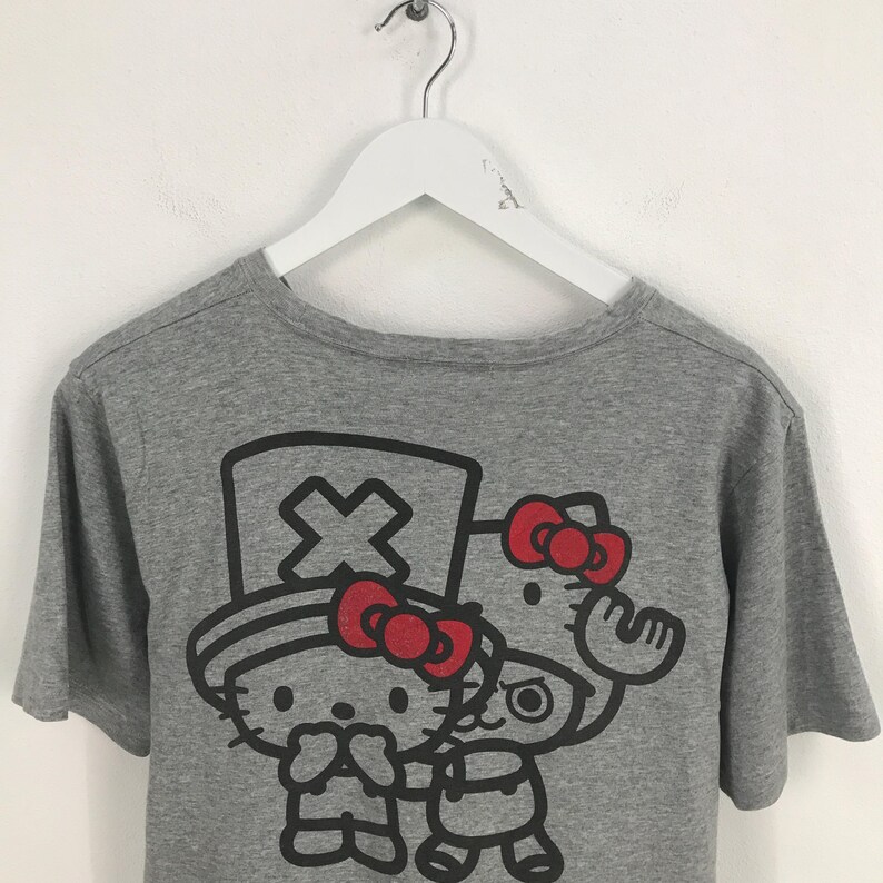 Pu&ograve; includere: T-shirt grigio con una grafica di due personaggi dei cartoni animati con fiocchi rossi. Un personaggio indossa un cappello con un simbolo a croce. L'altro personaggio ha un fiocco rosso e sta salutando. La maglietta &egrave; appesa a un gancio bianco.