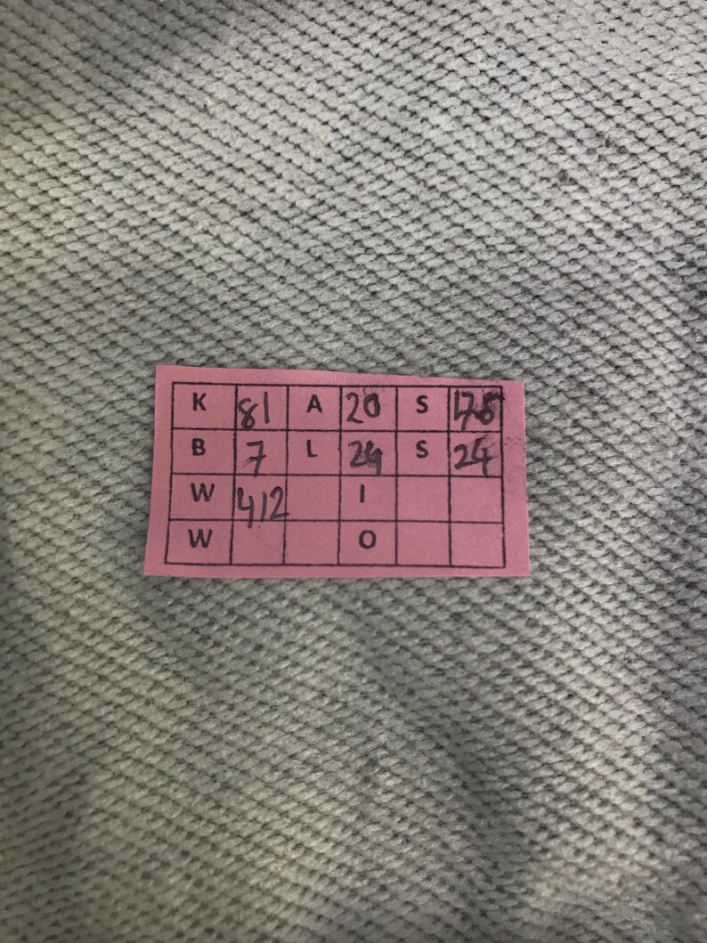 May include: A pink rectangular tag with black text and a grid pattern. The tag contains the letters K, B, W, A, L, S, and W, along with numbers. The background is a light grey, textured fabric.