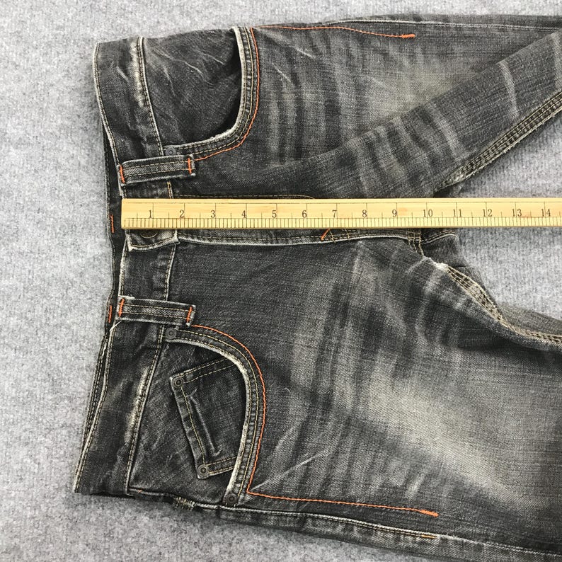 May include: A pair of black denim jeans with orange stitching. The jeans have a classic five-pocket design and a straight leg cut. A wooden ruler is placed across the jeans, measuring approximately 14 inches.