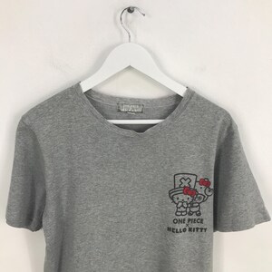 Pu&ograve; includere: Una maglietta grigia con una grafica di Hello Kitty a tema One Piece. La grafica mostra Hello Kitty con un cappello e un'arma. Il testo "ONE PIECE HELLO KITTY" &egrave; stampato sotto.