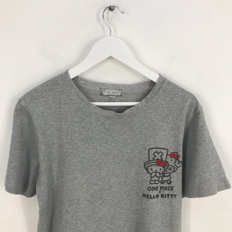 Pu&ograve; includere: Una maglietta grigia con una grafica di Hello Kitty a tema One Piece. La grafica mostra Hello Kitty con un cappello e un'arma. Il testo "ONE PIECE HELLO KITTY" &egrave; stampato sotto.