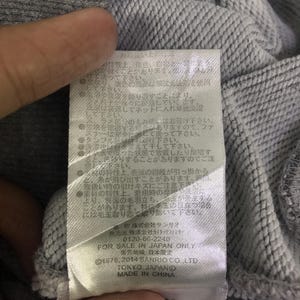 May include: A close-up shot of grey fabric with a label attached. The label features Japanese text and English text, including "FOR SALE IN JAPAN ONLY", "&copy;1976, 2014 SANRIO CO. LTD", "TOKYO JAPAND", and "MADE IN CHINA".