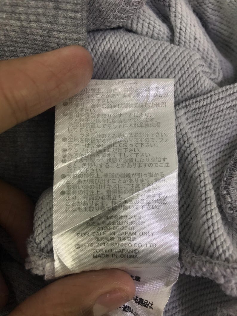 May include: A close-up shot of grey fabric with a label attached. The label features Japanese text and English text, including "FOR SALE IN JAPAN ONLY", "&copy;1976, 2014 SANRIO CO. LTD", "TOKYO JAPAND", and "MADE IN CHINA".