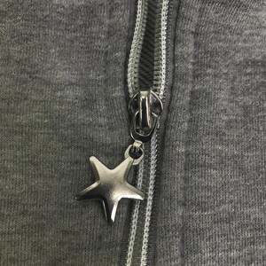 May include: Close-up of a grey zip-up hoodie with a silver star-shaped zipper pull. The zipper is silver and the fabric is a solid grey colour. The zipper pull is a five-pointed star.