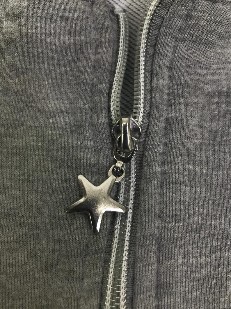May include: Close-up of a grey zip-up hoodie with a silver star-shaped zipper pull. The zipper is silver and the fabric is a solid grey colour. The zipper pull is a five-pointed star.