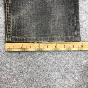 May include: Close-up of a section of dark gray denim jeans with a wooden ruler placed below. The ruler shows measurements in inches. The jeans have visible stitching along the side seam.