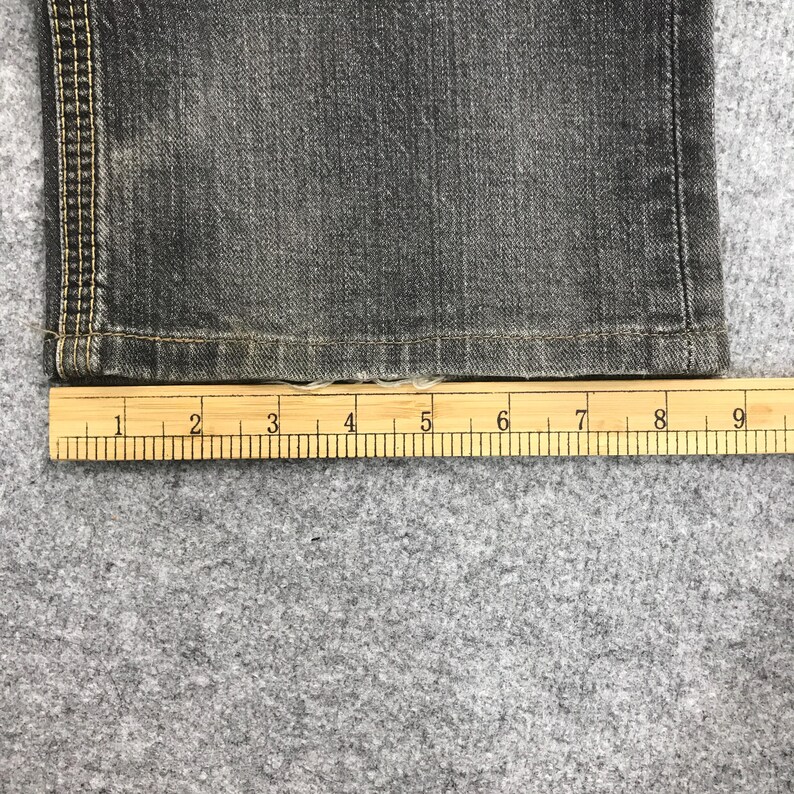 May include: Close-up of a section of dark gray denim jeans with a wooden ruler placed below. The ruler shows measurements in inches. The jeans have visible stitching along the side seam.
