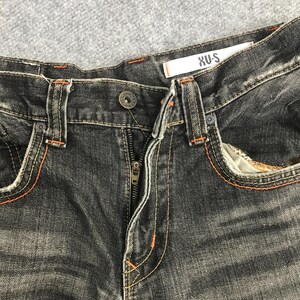 May include: Close-up of black denim jeans with orange stitching. The jeans have a button closure and a zipper. The brand name "XU-S" is visible on a white tag. The jeans have front pockets.