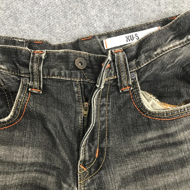 May include: Close-up of black denim jeans with orange stitching. The jeans have a button closure and a zipper. The brand name "XU-S" is visible on a white tag. The jeans have front pockets.