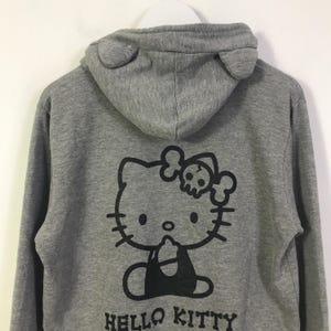 May include: Grey hooded sweatshirt with a black Hello Kitty graphic on the back, incorporating a skull and crossbones. The hood has ear-shaped details. The words "HELLO KITTY" are printed below the graphic.