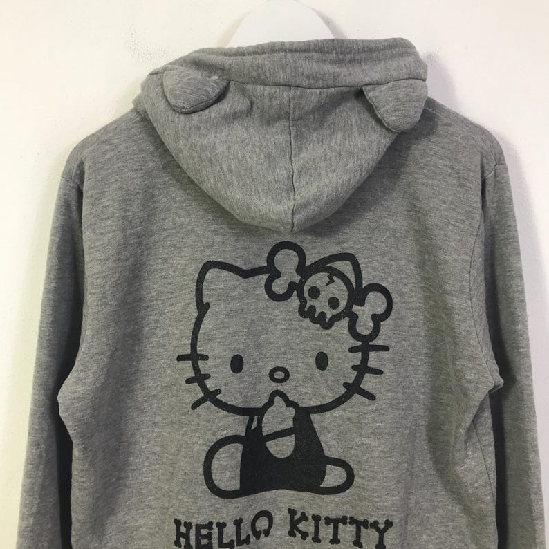 May include: Grey hooded sweatshirt with a black Hello Kitty graphic on the back, incorporating a skull and crossbones. The hood has ear-shaped details. The words "HELLO KITTY" are printed below the graphic.
