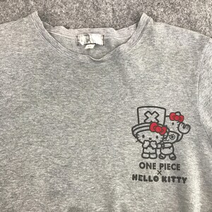 Pu&ograve; includere: Una maglietta girocollo grigio melange con una grafica di Hello Kitty e un personaggio di One Piece. La grafica include il testo "ONE PIECE x HELLO KITTY" in nero. La maglietta ha una piccola etichetta sul collo.