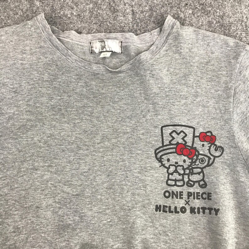Pu&ograve; includere: Una maglietta girocollo grigio melange con una grafica di Hello Kitty e un personaggio di One Piece. La grafica include il testo "ONE PIECE x HELLO KITTY" in nero. La maglietta ha una piccola etichetta sul collo.