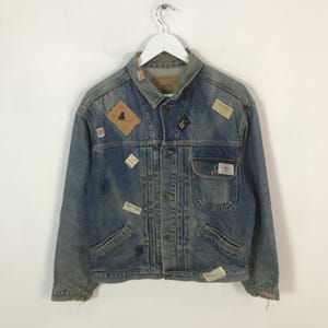 May include: A vintage denim jacket with multiple patches of various shapes and sizes. The jacket is a light blue wash with a button-up front, pockets, and a collar. Patches include geometric shapes and text.