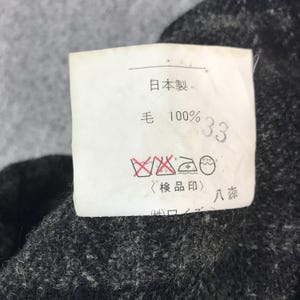 May include: A close-up shot of a white label with Japanese text and symbols, including "日本製" and "毛 100% 33". The label is attached to a dark grey, textured fabric.