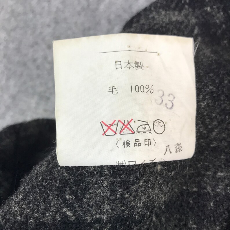 May include: A close-up shot of a white label with Japanese text and symbols, including "日本製" and "毛 100% 33". The label is attached to a dark grey, textured fabric.
