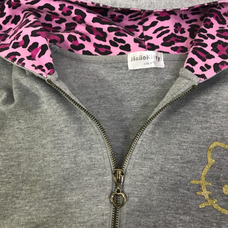May include: A gray zip-up hoodie with a pink and black leopard print lining in the hood. The zipper has a heart-shaped pull. The Hello Kitty logo is in gold on the lower right. The label reads "Hello Kitty -3L-".
