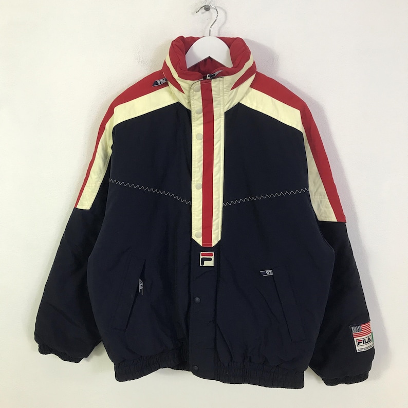 May include: A vintage navy blue and red Fila jacket with a cream stripe design. The jacket features a high collar, zip pockets, and the Fila logo. The jacket has a USA flag patch on the sleeve.