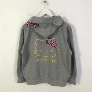 May include: A gray zip-up hoodie featuring a gold Hello Kitty design on the back. The design includes a bow with pink and black leopard print. The hood has ears and a pink leopard print lining. The text "Hello Kitty" is below the design.