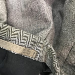 May include: Close-up of grey herringbone fabric with a black and white pattern. The fabric is folded, revealing a glimpse of a black lining and a beige waistband. The texture of the fabric is visible, showcasing the detailed weave.